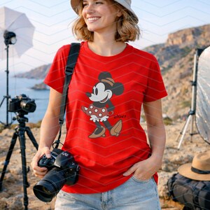 K&ouml;nnte beinhalten: Ein rotes T-Shirt mit einem Minnie Mouse-Motiv. Die Zeichentrickfigur ist in einer klassischen Pose dargestellt und tr&auml;gt ein gepunktetes Kleid und einen Hut. Das T-Shirt wird mit einer blauen Jeans kombiniert. Im Hintergrund sind Fotoausr&uuml;stung und eine Kamera zu sehen.