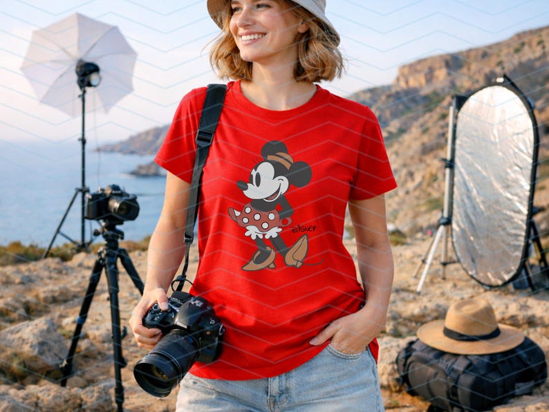 K&ouml;nnte beinhalten: Ein rotes T-Shirt mit einem Minnie Mouse-Motiv. Die Zeichentrickfigur ist in einer klassischen Pose dargestellt und tr&auml;gt ein gepunktetes Kleid und einen Hut. Das T-Shirt wird mit einer blauen Jeans kombiniert. Im Hintergrund sind Fotoausr&uuml;stung und eine Kamera zu sehen.