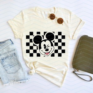 May include: A white t-shirt with a black and white checkered pattern and a black and white cartoon image of Mickey Mouse winking. The Mickey Mouse image is in the center of the checkered pattern.