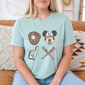 May include: A light blue t-shirt with a Mickey Mouse graphic wearing a baseball cap with the letter "M" on it. The graphic also includes a baseball glove, a baseball, and two crossed baseball bats. The text "#1" is on the glove.