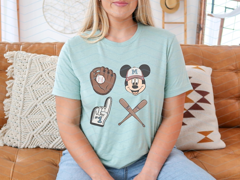 May include: A light blue t-shirt with a Mickey Mouse graphic wearing a baseball cap with the letter "M" on it. The graphic also includes a baseball glove, a baseball, and two crossed baseball bats. The text "#1" is on the glove.