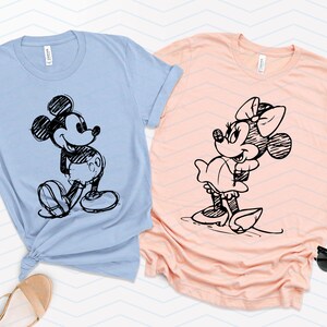 以下が含まれることがあります： 薄い青と桃色のTシャツ2枚。黒と白のスケッチでミッキーマウスとミニーマウスが描かれている。