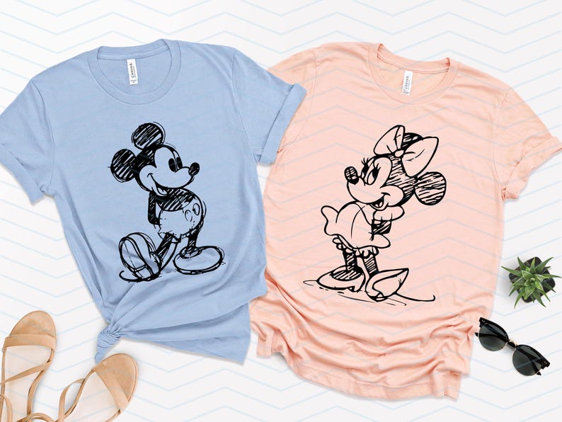 以下が含まれることがあります： 薄い青と桃色のTシャツ2枚。黒と白のスケッチでミッキーマウスとミニーマウスが描かれている。
