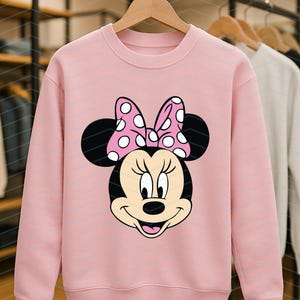 May include: A light pink sweatshirt featuring a graphic of Minnie Mouse's face. Minnie's face is black and tan with a pink bow with white polka dots. The sweatshirt is hanging on a wooden hanger.
