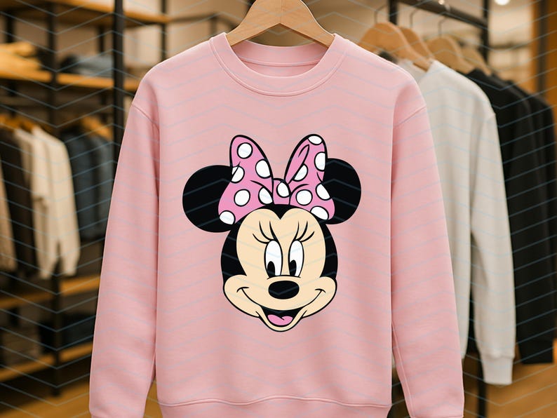 May include: A light pink sweatshirt featuring a graphic of Minnie Mouse's face. Minnie's face is black and tan with a pink bow with white polka dots. The sweatshirt is hanging on a wooden hanger.
