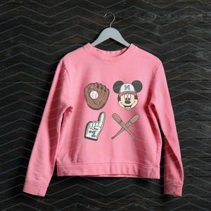 May include: Pink sweatshirt with a Mickey Mouse baseball graphic. The graphic includes a baseball glove, a baseball bat, and a number one finger. The Mickey Mouse character is wearing a baseball cap with the letter "M" on it.