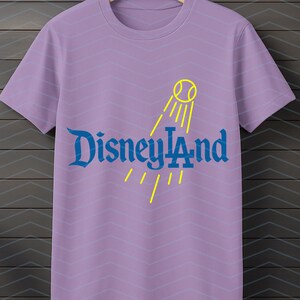May include: Lavender t-shirt with the text "Disneyland" in blue, and a yellow tennis ball and sun ray design. The shirt has a crew neck and short sleeves. The background has a chevron pattern.