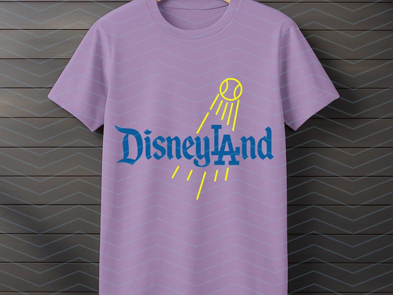 May include: Lavender t-shirt with the text "Disneyland" in blue, and a yellow tennis ball and sun ray design. The shirt has a crew neck and short sleeves. The background has a chevron pattern.