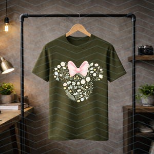 May include: Olive green t-shirt featuring a floral design in the shape of a mouse head with a pink bow. The design includes white and yellow flowers and green leaves. The t-shirt is hanging on a black clothing rack.