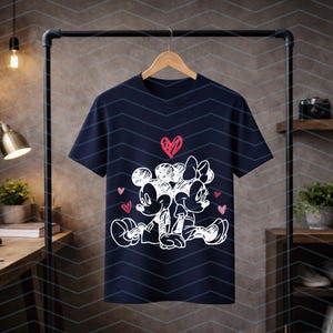 Peut inclure: T-shirt bleu marine avec un dessin au trait blanc de Mickey et Minnie Mouse, entour&eacute;s de coeurs roses. Le tee-shirt est pr&eacute;sent&eacute; sur un portant noir. Le design est de style dessin anim&eacute;.