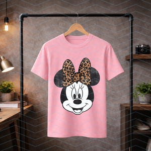 May include: A pink t-shirt featuring a Minnie Mouse graphic. The graphic shows Minnie's face with a black head and a bow with a leopard print design. The t-shirt is hanging on a wooden hanger.