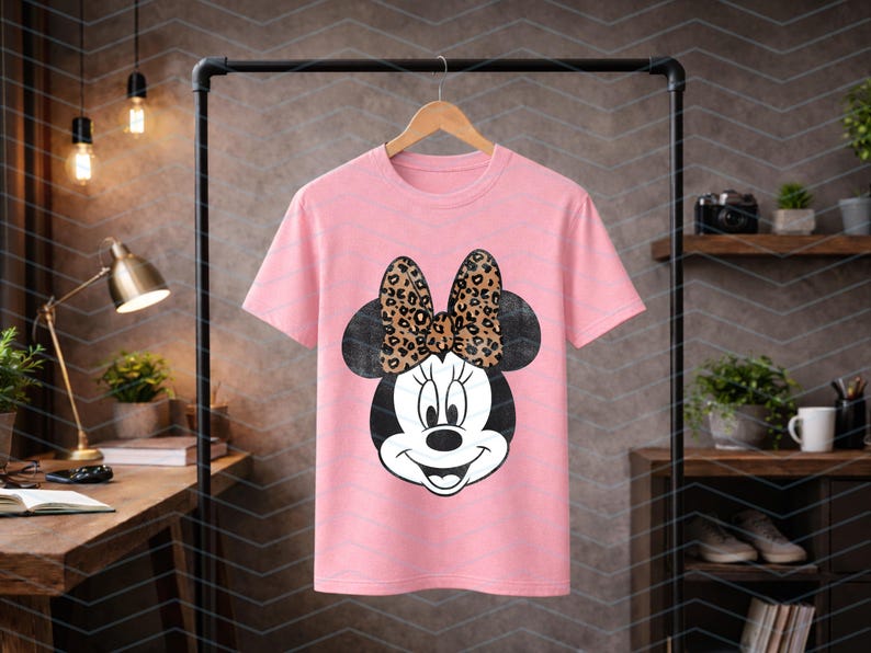 May include: A pink t-shirt featuring a Minnie Mouse graphic. The graphic shows Minnie's face with a black head and a bow with a leopard print design. The t-shirt is hanging on a wooden hanger.