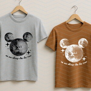 Puede incluir: Dos camisetas con un gr&aacute;fico de la cabeza de Mickey Mouse hecha de bolas de discoteca. El texto en las camisetas dice "Me and Mickey vibe like that". Una camiseta es gris y la otra es marr&oacute;n.