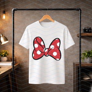 Puede incluir: Camiseta blanca con un gran lazo rojo con lunares blancos. El lazo tiene una textura brillante y un contorno negro. La camiseta est&aacute; colgada en una percha de madera.
