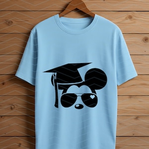 May include: Light blue t-shirt featuring a black graphic of a mouse wearing a graduation cap and sunglasses with a heart-shaped detail. The shirt is hanging on a wooden hanger against a wood-paneled background.