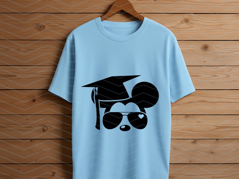 May include: Light blue t-shirt featuring a black graphic of a mouse wearing a graduation cap and sunglasses with a heart-shaped detail. The shirt is hanging on a wooden hanger against a wood-paneled background.