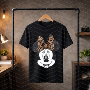May include: Black t-shirt featuring a Minnie Mouse graphic. The graphic shows Minnie's face with a large bow in a leopard print pattern. The t-shirt is hanging on a wooden hanger.