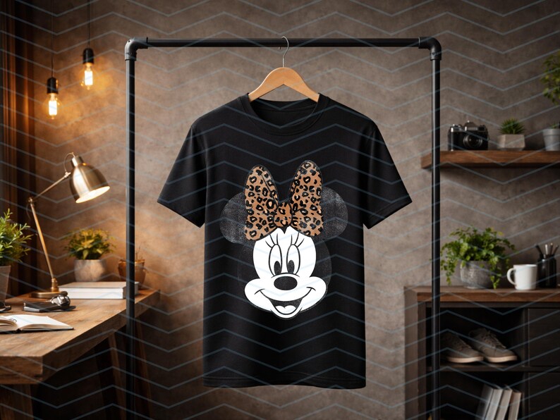 May include: Black t-shirt featuring a Minnie Mouse graphic. The graphic shows Minnie's face with a large bow in a leopard print pattern. The t-shirt is hanging on a wooden hanger.