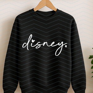 Op de afbeelding: Zwarte crewneck sweatshirt met het woord "disney" in witte letters. De "i" in het woord "disney" is vervangen door een Mickey Mouse-hoofd. De sweatshirt heeft een chevron patroon.
