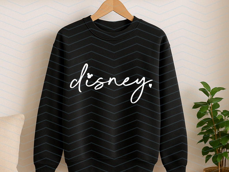 Op de afbeelding: Zwarte crewneck sweatshirt met het woord "disney" in witte letters. De "i" in het woord "disney" is vervangen door een Mickey Mouse-hoofd. De sweatshirt heeft een chevron patroon.
