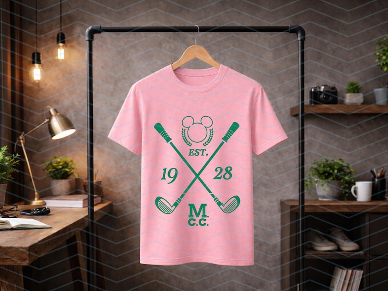May include: A light pink t-shirt with a green golf-themed design. The design includes crossed golf clubs, the text "EST. 1928 M.C.C.", and a Mickey Mouse-inspired emblem. The shirt is hanging on a black clothing rack.