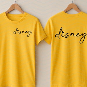 Op de afbeelding: Twee gele t-shirts met het woord "disney" in zwart script. E&eacute;n shirt heeft de tekst op de voorkant, de andere op de achterkant. Het woord "disney" heeft een hartje boven de "i".