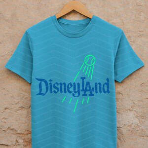 May include: A turquoise t-shirt with a chevron pattern. The shirt features the text "Disneyland" in a stylized font, with a green tennis ball graphic and lines. The shirt is displayed on a wooden hanger.