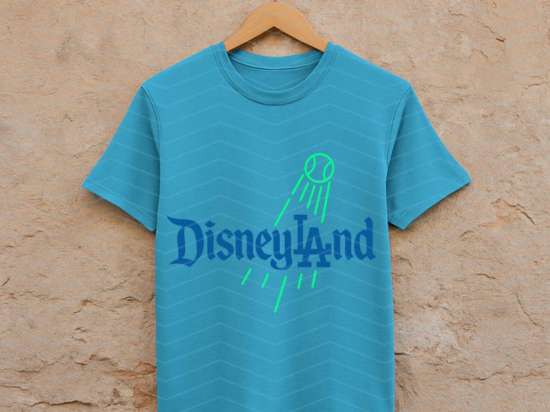 May include: A turquoise t-shirt with a chevron pattern. The shirt features the text "Disneyland" in a stylized font, with a green tennis ball graphic and lines. The shirt is displayed on a wooden hanger.