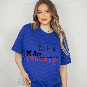 Op de afbeelding: Een koninklijk blauw T-shirt met de tekst "I'm Her Mickey" in zwarte en rode letters. Het Mickey Mouse-hoofd is zwart omlijnd en rood gevuld.