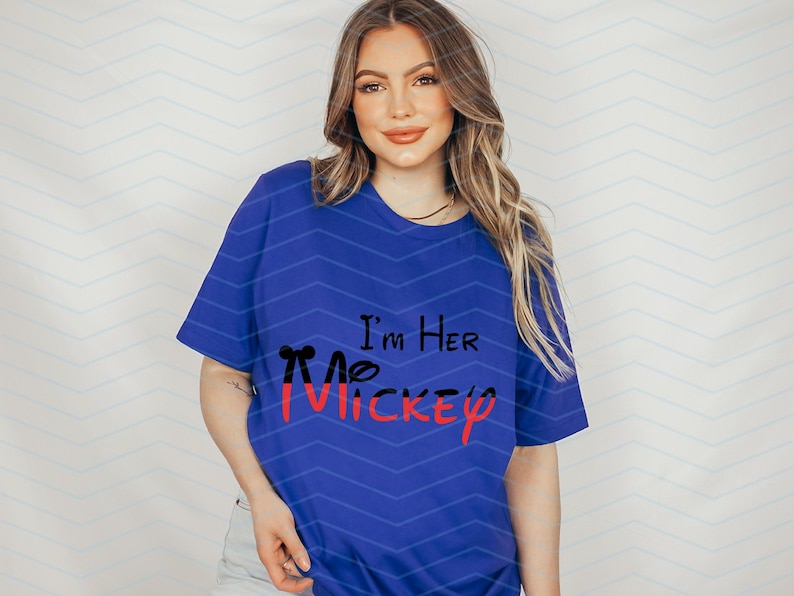 Op de afbeelding: Een koninklijk blauw T-shirt met de tekst "I'm Her Mickey" in zwarte en rode letters. Het Mickey Mouse-hoofd is zwart omlijnd en rood gevuld.
