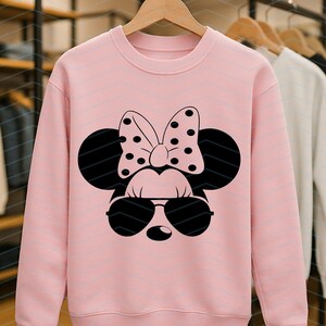 May include: A light pink sweatshirt featuring a black graphic of a cartoon mouse face with sunglasses and a polka-dot bow. The sweatshirt is hanging on a wooden hanger in a clothing store.