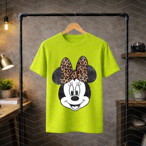 May include: A lime green t-shirt featuring a Minnie Mouse graphic. The graphic shows Minnie's face with a black head and ears, a white face, and a bow with a leopard print design. The shirt is hanging on a wooden hanger.
