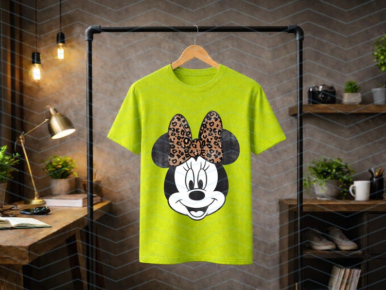 May include: A lime green t-shirt featuring a Minnie Mouse graphic. The graphic shows Minnie's face with a black head and ears, a white face, and a bow with a leopard print design. The shirt is hanging on a wooden hanger.