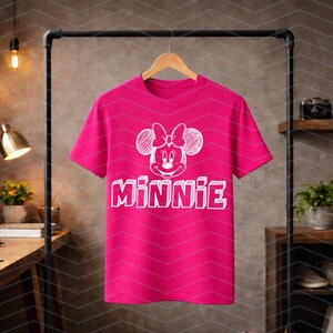 May include: A vibrant pink t-shirt displaying a white Minnie Mouse graphic with a bow and the name "MINNIE". The tee is suspended on a wooden hanger. The background is a neutral colour.