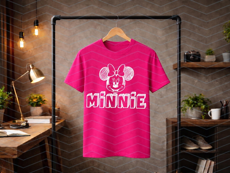 May include: A vibrant pink t-shirt displaying a white Minnie Mouse graphic with a bow and the name "MINNIE". The tee is suspended on a wooden hanger. The background is a neutral colour.