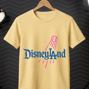 May include: A pale yellow t-shirt featuring the blue text "Disneyland" and a pink baseball graphic. The shirt is displayed on a wooden hanger. The background is dark.