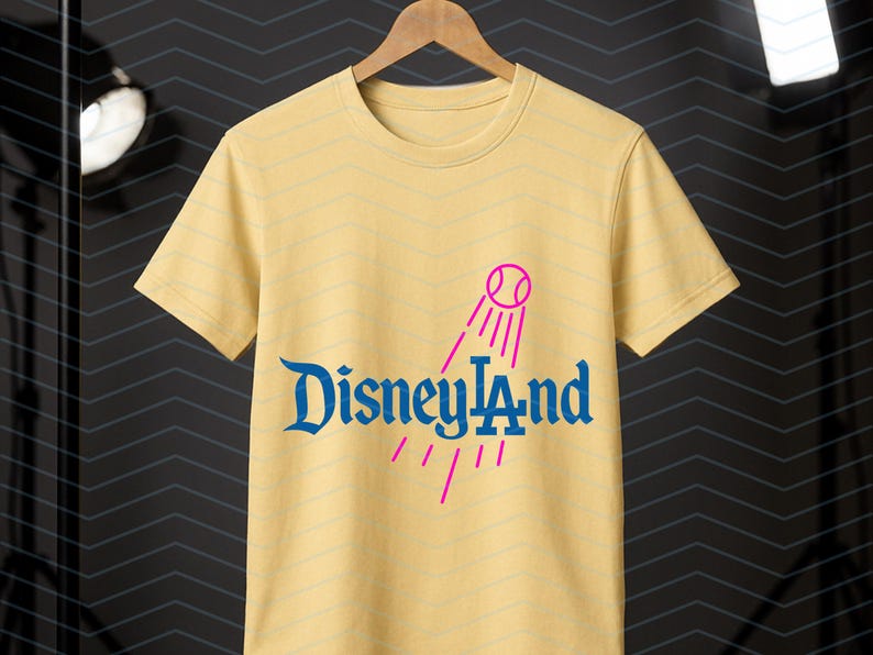 May include: A pale yellow t-shirt featuring the blue text "Disneyland" and a pink baseball graphic. The shirt is displayed on a wooden hanger. The background is dark.