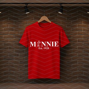 Pu&ograve; includere: Una maglietta rossa con la scritta "Minnie" in bianco, una grafica e il testo "Est. 1928". La maglietta &egrave; appesa a una gruccia di legno su uno sfondo di muro di mattoni.