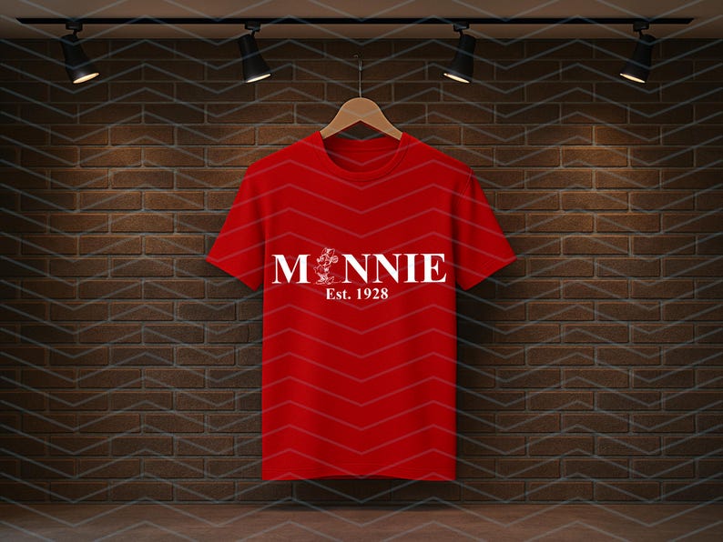 Pu&ograve; includere: Una maglietta rossa con la scritta "Minnie" in bianco, una grafica e il testo "Est. 1928". La maglietta &egrave; appesa a una gruccia di legno su uno sfondo di muro di mattoni.