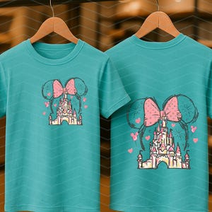 Pu&ograve; includere: Due magliette color turchese con un fiocco rosa e un'immagine di cartone animato del castello di Cenerentola. Il castello &egrave; circondato da cuori rosa e fuochi d'artificio.