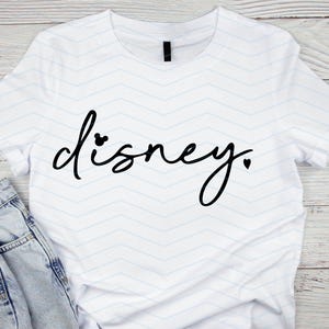 Op de afbeelding: Wit T-shirt met het woord "Disney" in zwart script. De "i" in Disney is gestippeld met een Mickey Mouse-hoofd, en een hart vervangt de punt. Een blauwe jeans is zichtbaar.