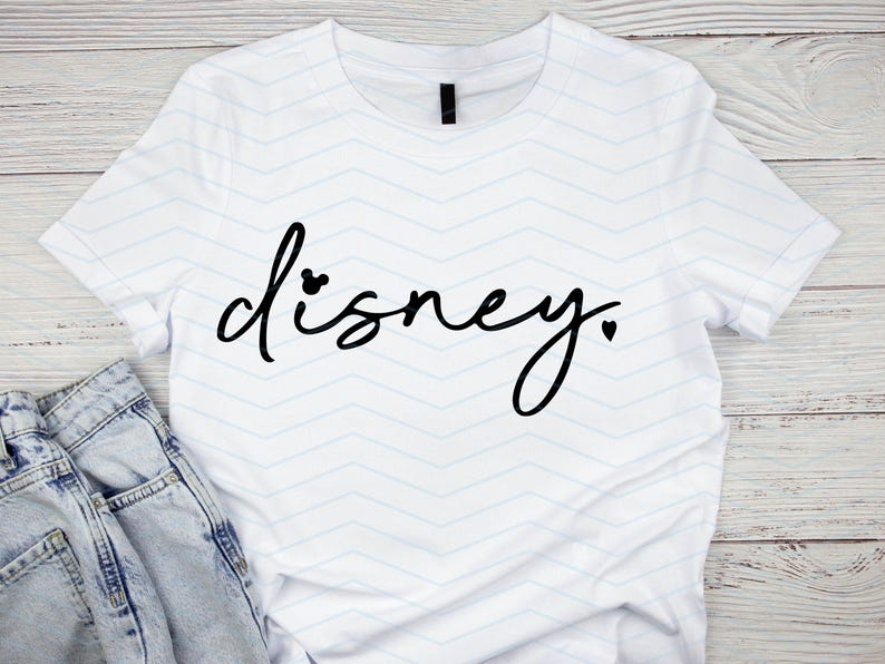 Op de afbeelding: Wit T-shirt met het woord "Disney" in zwart script. De "i" in Disney is gestippeld met een Mickey Mouse-hoofd, en een hart vervangt de punt. Een blauwe jeans is zichtbaar.
