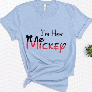 Op de afbeelding: Lichtblauw T-shirt met de tekst "I'm Her Mickey" in zwarte en rode letters. De Mickey is gestileerd met een zwarte omtrek en rode vulling.