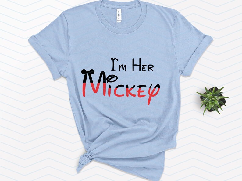 Op de afbeelding: Lichtblauw T-shirt met de tekst "I'm Her Mickey" in zwarte en rode letters. De Mickey is gestileerd met een zwarte omtrek en rode vulling.