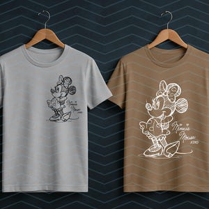 May include: Two t-shirts, one gray and one brown, each featuring a sketch of Minnie Mouse. The Minnie Mouse design includes a bow, dress, and the words "Minnie Mouse xoxo". The shirts are hanging on wooden hangers.