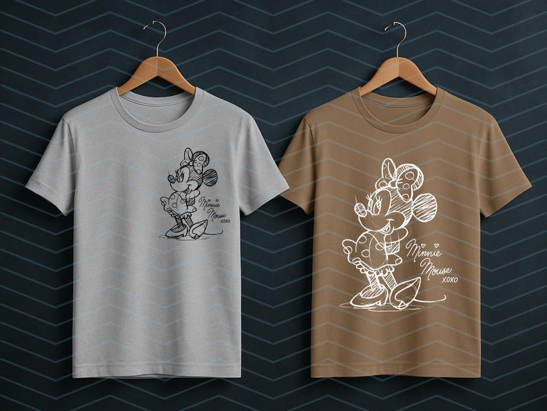 May include: Two t-shirts, one gray and one brown, each featuring a sketch of Minnie Mouse. The Minnie Mouse design includes a bow, dress, and the words "Minnie Mouse xoxo". The shirts are hanging on wooden hangers.
