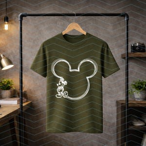 Puede incluir: Camiseta verde oliva con un contorno blanco de la cabeza y el cuerpo de Mickey Mouse. El dise&ntilde;o es una imagen de dibujos animados sencilla. La camiseta est&aacute; colgada en una percha negra.