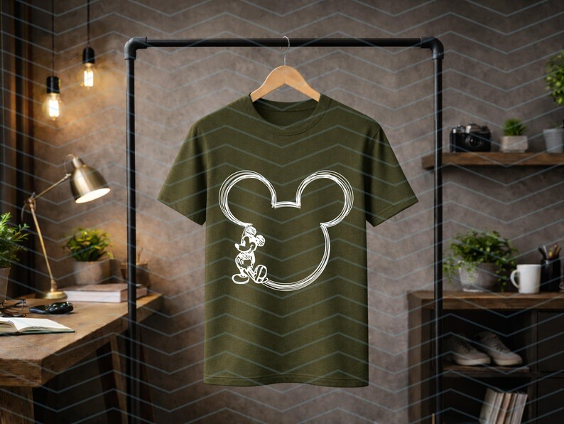 Puede incluir: Camiseta verde oliva con un contorno blanco de la cabeza y el cuerpo de Mickey Mouse. El dise&ntilde;o es una imagen de dibujos animados sencilla. La camiseta est&aacute; colgada en una percha negra.