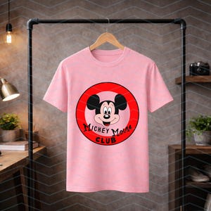 May include: A light pink short-sleeved t-shirt featuring a circular graphic on the chest. The graphic contains a red border, a central illustration of Mickey Mouse, and the black text "Mickey Mouse Club" printed in a stylized font below the face.