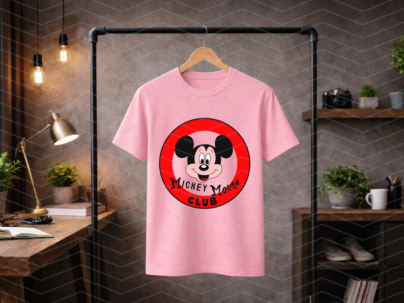 May include: A light pink short-sleeved t-shirt featuring a circular graphic on the chest. The graphic contains a red border, a central illustration of Mickey Mouse, and the black text "Mickey Mouse Club" printed in a stylized font below the face.